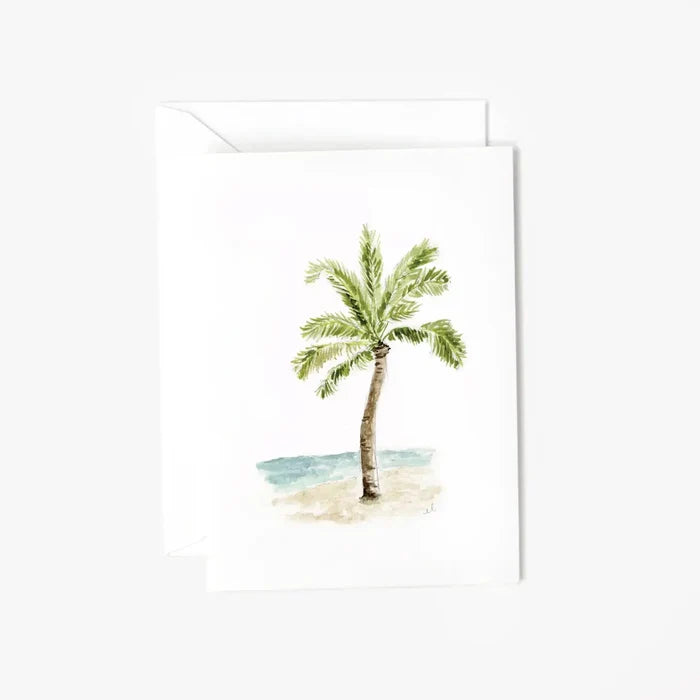 Watercolor palm tree greeting card featuring a hand-painted palm tree on a sandy beach with soft blue ocean waves, printed on a white folded card with matching envelope.