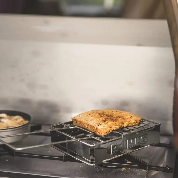 Primus toaster being used with a portable camping gas stove to make toast on a blurred background