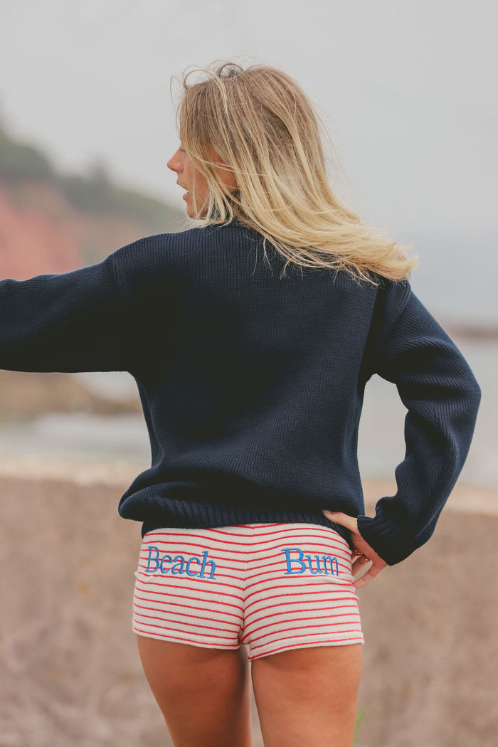 A woman standing on a beach, viewed from behind, wearing a dark navy knit sweater and pink and white striped shorts with the words 'Beach Bum' embroidered in blue.