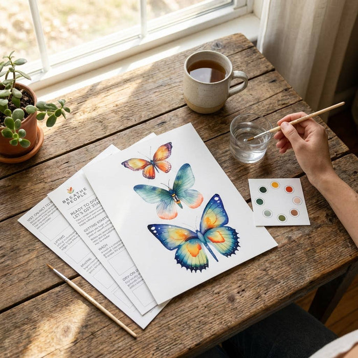 Person painting butterflies on paper with a cup of tea and art supplies on a wooden table.