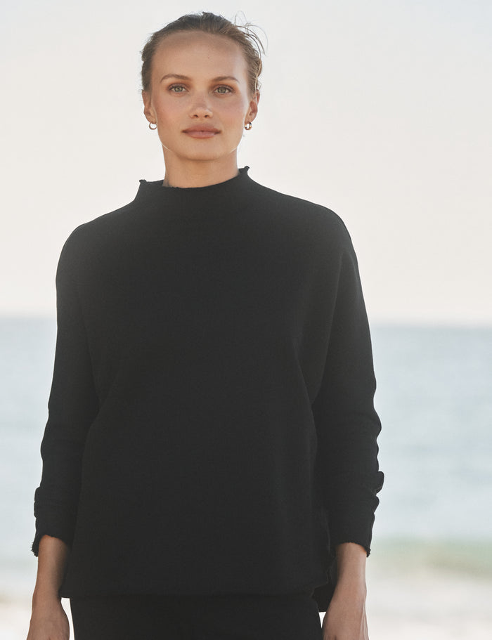 Woman wearing a black sweater standing on a beach with ocean in the background