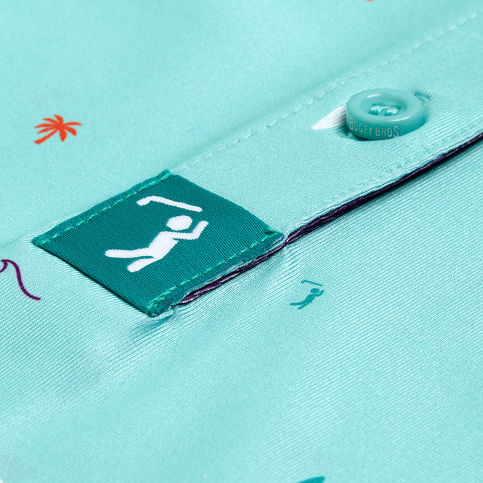 Close-up of the Beach Women's Polo, an athletic fit teal shirt with moisture-wicking fabric, featuring a button and small prints of palm trees and swimmers, plus a swimmer icon on the tag.