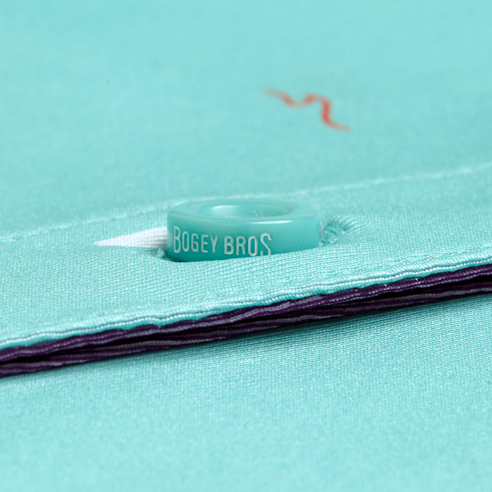 Close-up of a light teal button engraved with BOGEY BROS, sewn onto moisture-wicking teal fabric with purple lining on the Beach Women's Polo. A hint of white and a red embroidered line appear in the background, highlighting the button and stitching.