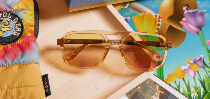 Camp Eyewear Grateful Dead Forever sunglasses with champagne translucent frames and gradient yellow-to-pink polarized lenses styled beside colorful artwork and accessories.