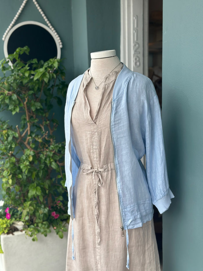 Light blue linen zip-front jacket with ribbed collar and cuffs, styled over a neutral linen dress. Features a relaxed fit, lightweight fabric, side pockets, and adjustable drawstring hem details.