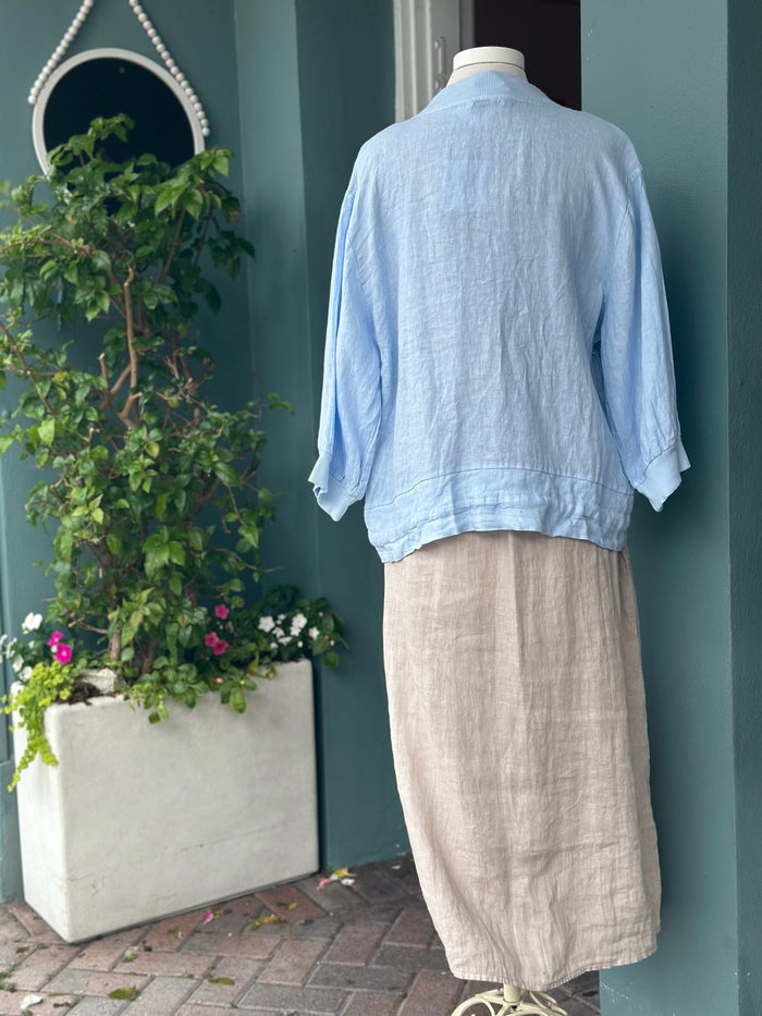 Back view of a light blue linen jacket with a relaxed silhouette, ribbed collar and cuffs, and a slightly cropped hem. Styled over a neutral linen dress.