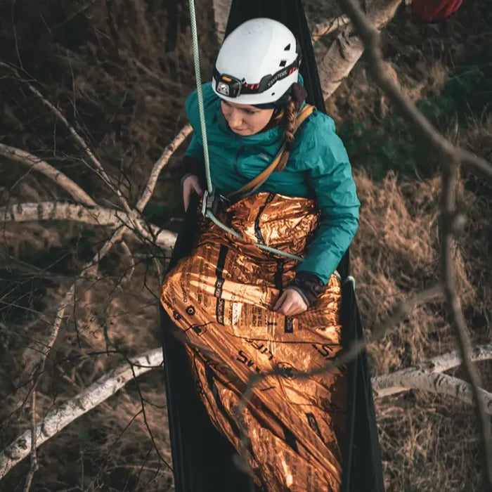 A person in a white helmet and teal jacket sits in a black hammock outdoors, wrapped in a Lifesystems heatshield thermal blanket