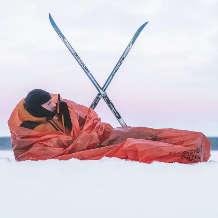 A person lying in snow wrapped in a bright orange Lifesystems Plastic Survival Bag, with crossed skis behind them against a pale sky.