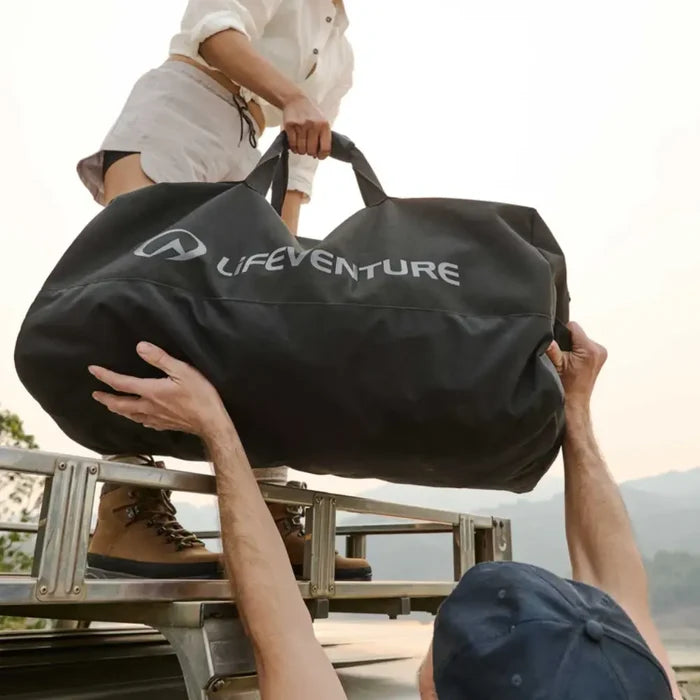 Two people lifting a Lifeventure Expedition Duffle Bag on a vehicle's roof rack with a scenic background.