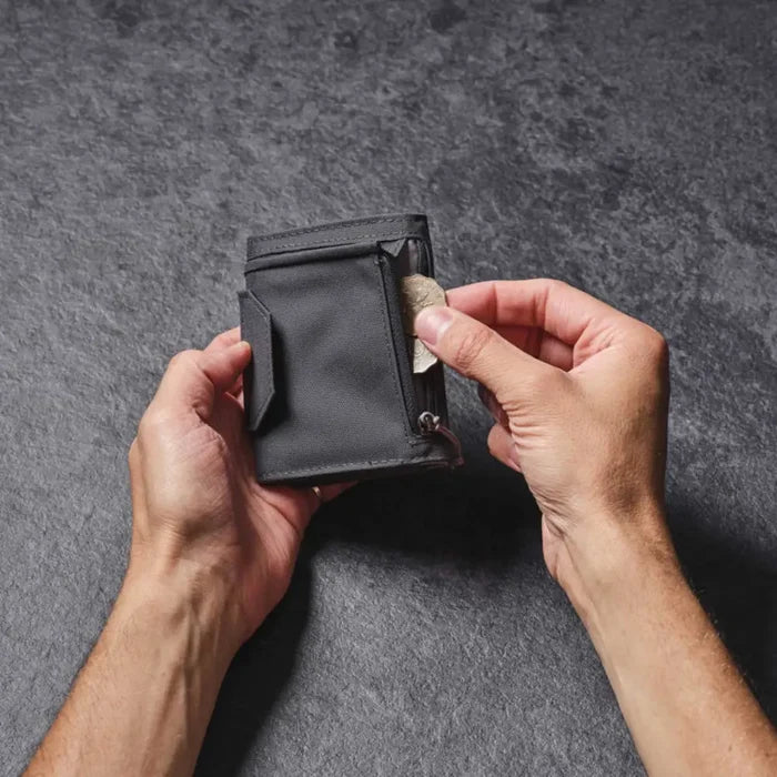 Person holding a Lifeventure RFiD Tri-Fold Wallet wit hthe right hand placing coins in the external zipped pocket, on a grey background