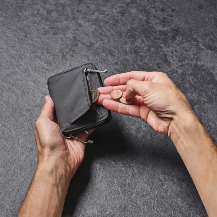 Person placing  coins into the external zipped pocket of a Lifeventure RFiD Zipped Bi-Fold Wallet, on a grey background