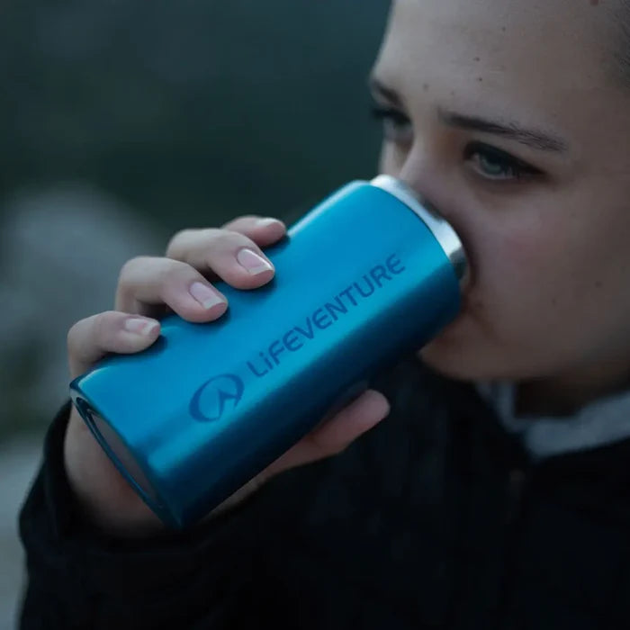 Person drinking from a blue Lifeventure stainless steel thermal mug outdoors.