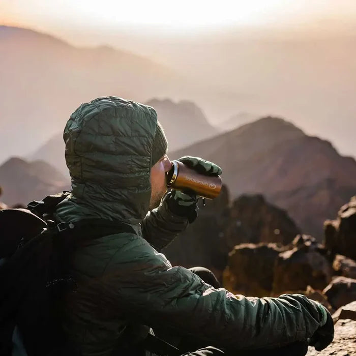 Person in a green jacket sitting on a mountain, drinking frm a Lifeventure Stainless Steel Thermal Mugin copper colour