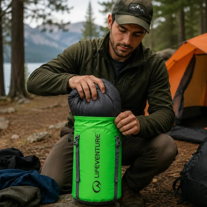 Man stuffing his sleeping bag into a Lifeventure Ultralight Compression Sack 15 Litres in green whilst camping in a forest by a lake