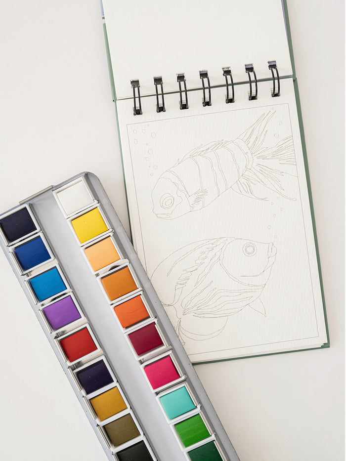Set of watercolor pans next to a coloring book with a fish design on a white background