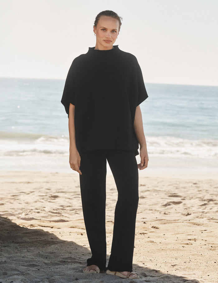 Person wearing a black capelet and pants standing on a beach with ocean in the background