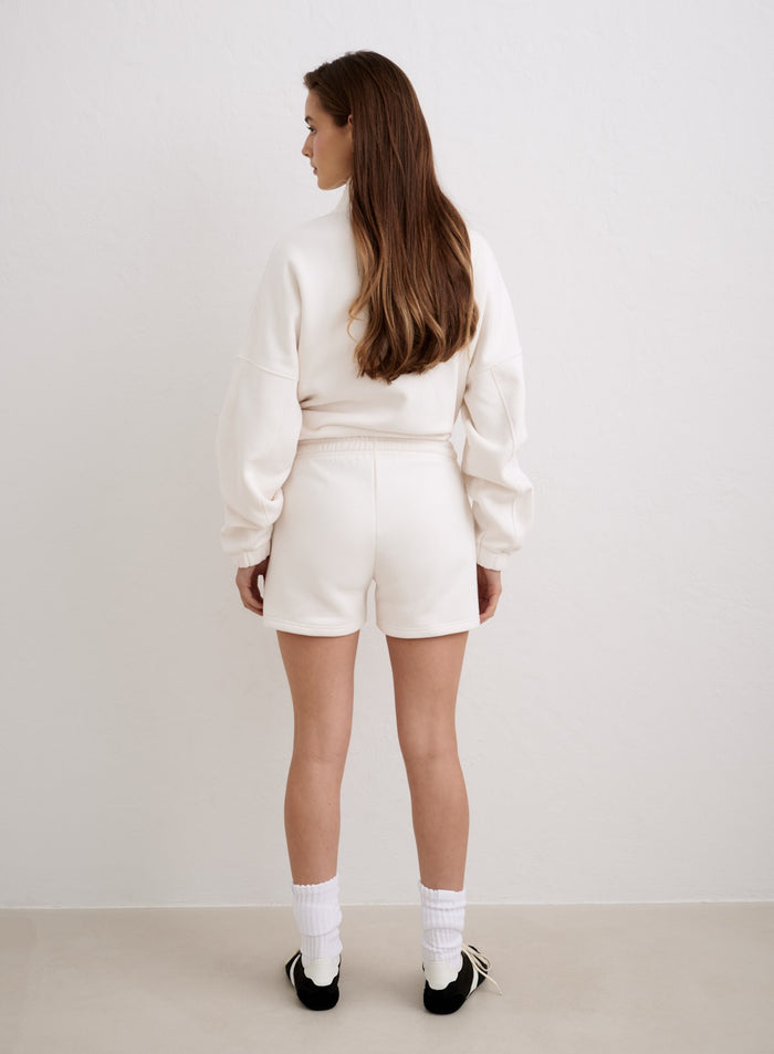 Nainen takaapäin yllään luonnonvalkoiset aim'n Script Sweat Shorts -collegeshortsit ja niihin sopiva collegepaita. [XS-174cm]