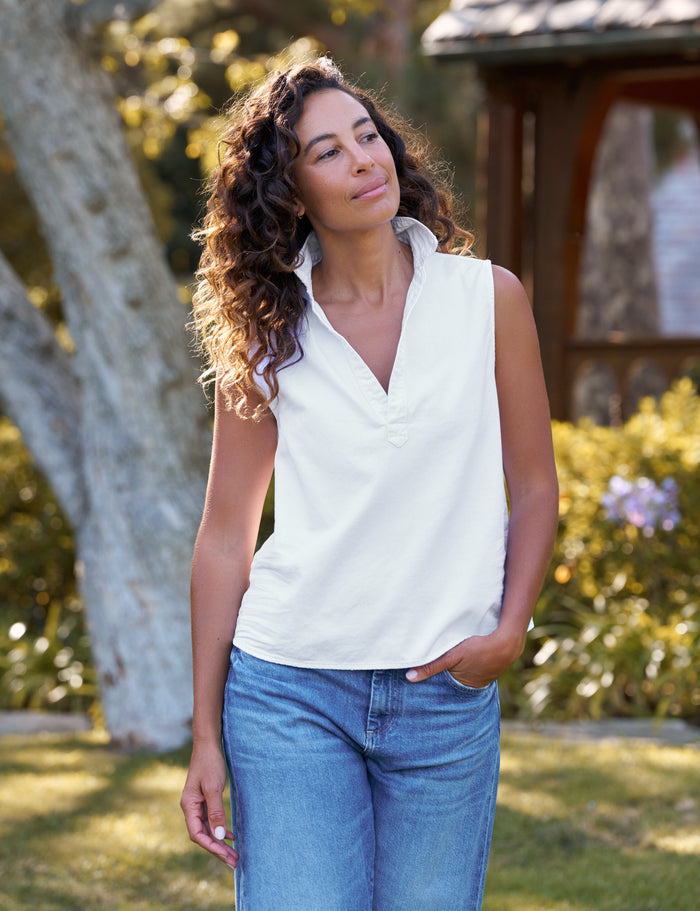 A woman with curly brown hair wearing a white sleeveless V-neck top and blue jeans, standing outdoors in a sunny garden setting.