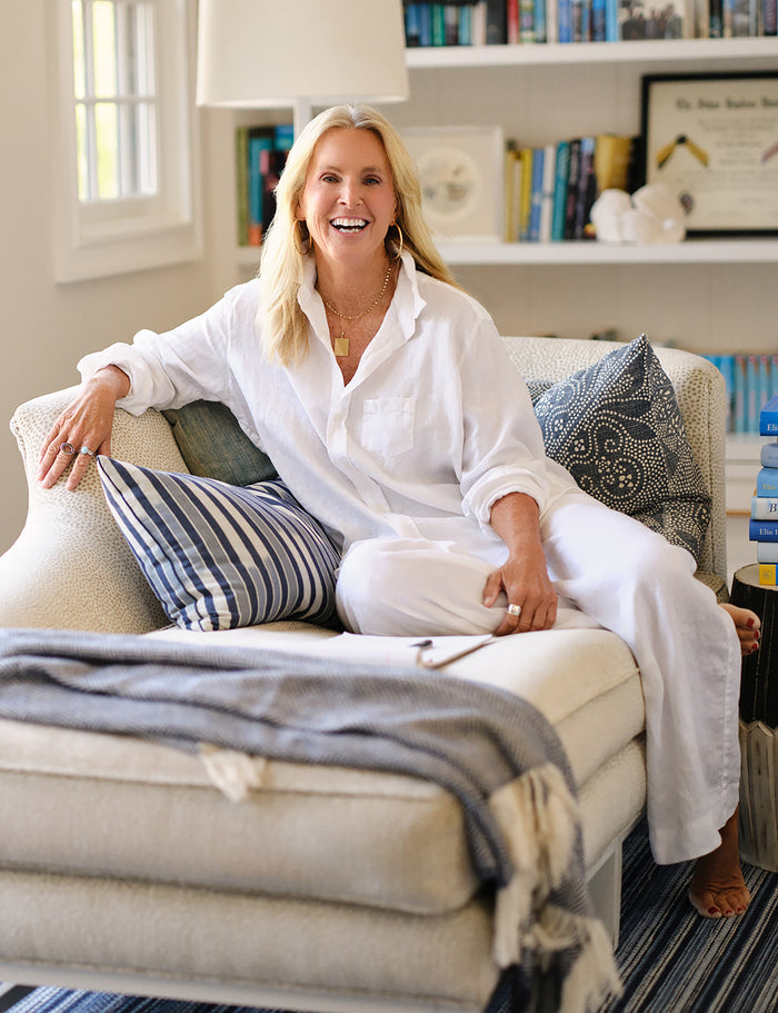A smiling blonde woman sits on a light beige sofa in a bright living room, wearing a loose white button-up shirt and white pants. The sofa has blue and white striped pillows and a gray throw blanket.