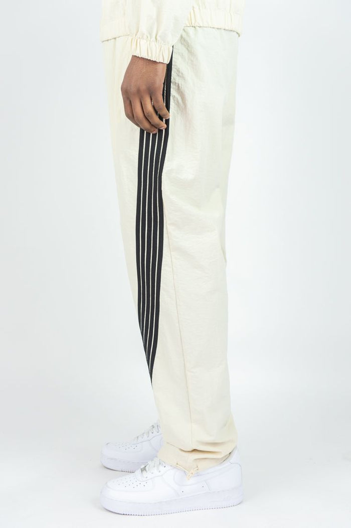 Cream Rebel Minds 'Bent Stripes' nylon sweatpants with relaxed fit and side stripes, shown on a model.