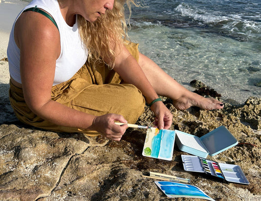Person sitting by the ocean, painting with watercolor on a rock