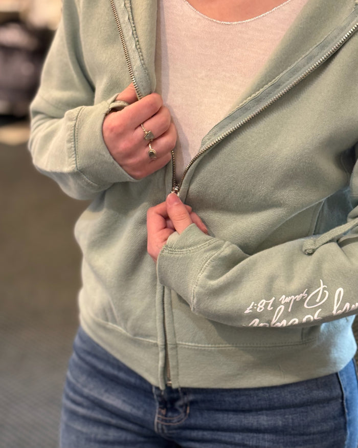 Woman wearing a sage green hoodie, handmade rings holding the zipper to show zip.