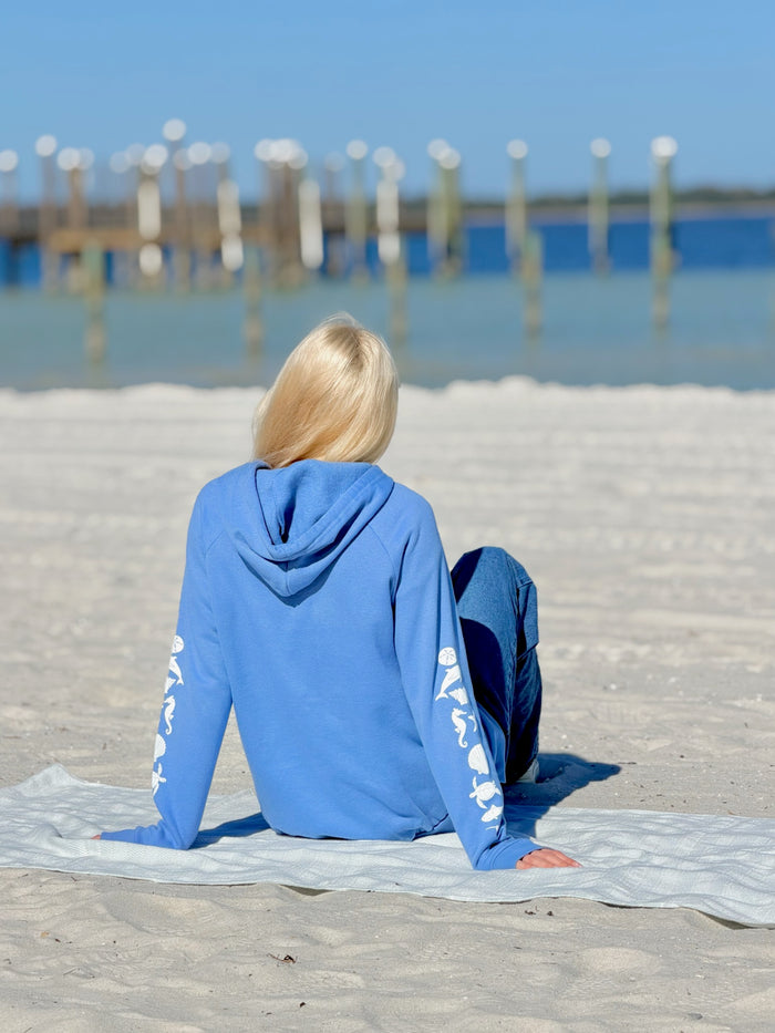 Person wearing a blue hoodie with sea life  on sleeves sitting on a towel at the beach.