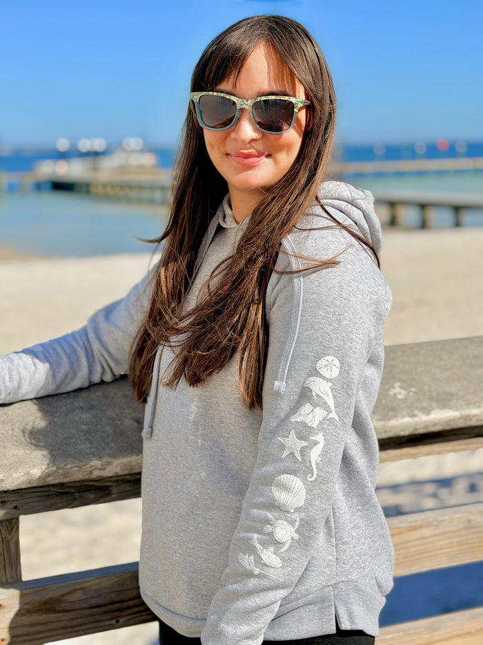 Woman wearing a gray hoodie with beach-themed designs by the ocean.