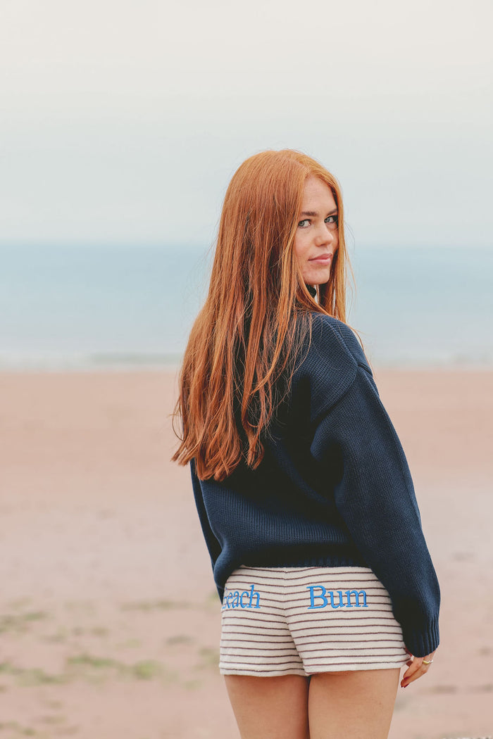 A woman with long red hair stands on a sandy beach looking back over her shoulder. She is wearing a dark navy blue knit sweater and cream-colored shorts with thin black stripes and blue text that reads 'Beach Bum'.