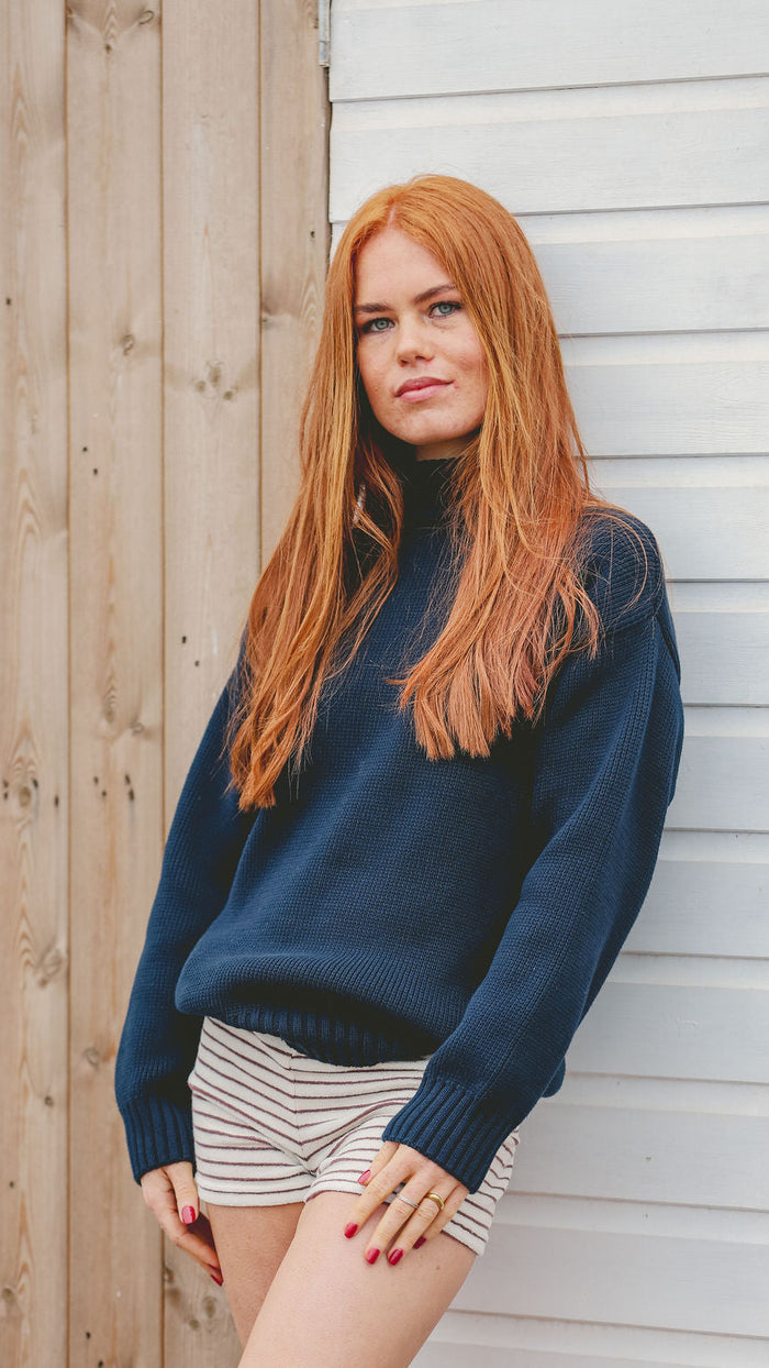 A woman with long red hair leans against a white paneled wall next to a wooden wall, wearing a dark navy blue turtleneck sweater and white shorts with thin black stripes.