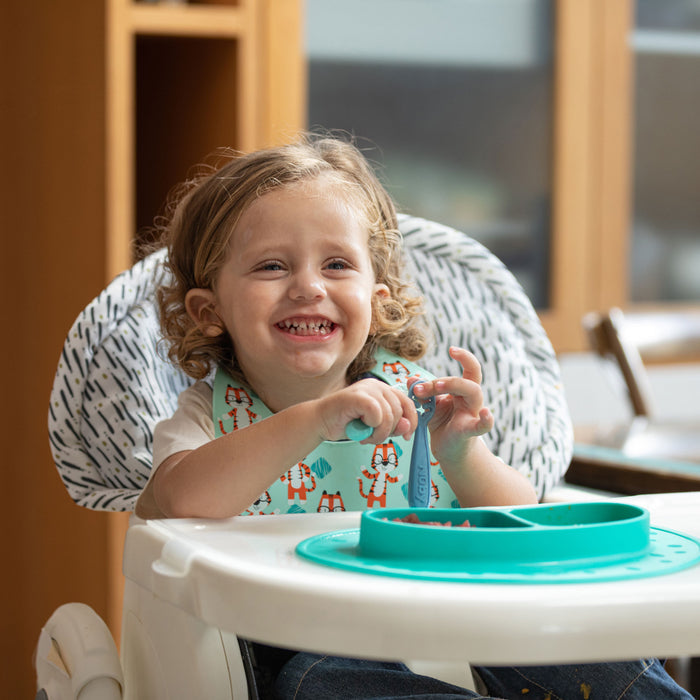 A smiling toddler with curly hair sits in a high chair, protected by the Nuby Water-Resistant High Chair Cover, wearing a bib with foxes and eating from a sectioned turquoise plate while holding a blue fork.