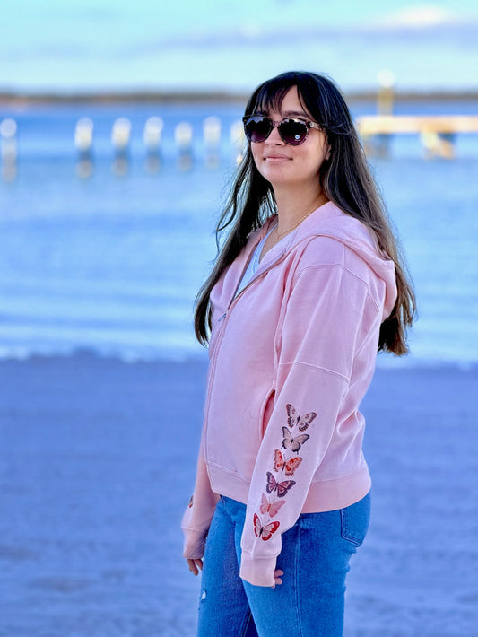 Woman wearing a pink hoodie with butterfly designs on sleeves at a beach.