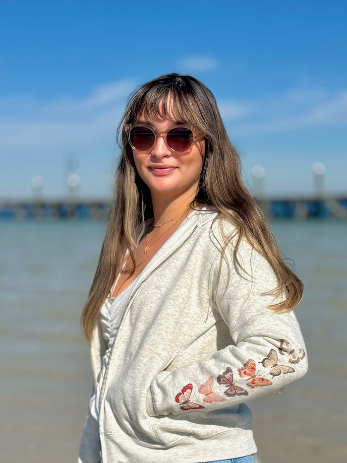 Woman wearing an oatmeal zip up with pink red butterfly designs on a beach.