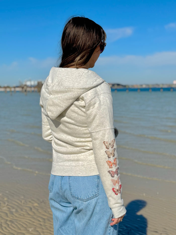 Person wearing a light-colored hoodie with butterfly designs on a beach