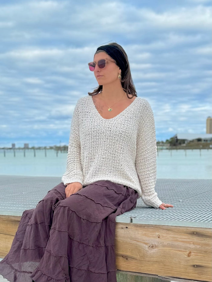Woman sitting by a waterfront wearing an ivory sweater and brown skirt with sunglasses on.