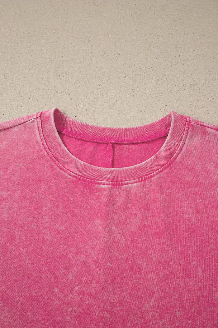 A close-up of a bright pink crewneck sweatshirt with a washed, mineral-washed texture, laid flat against a neutral beige background.